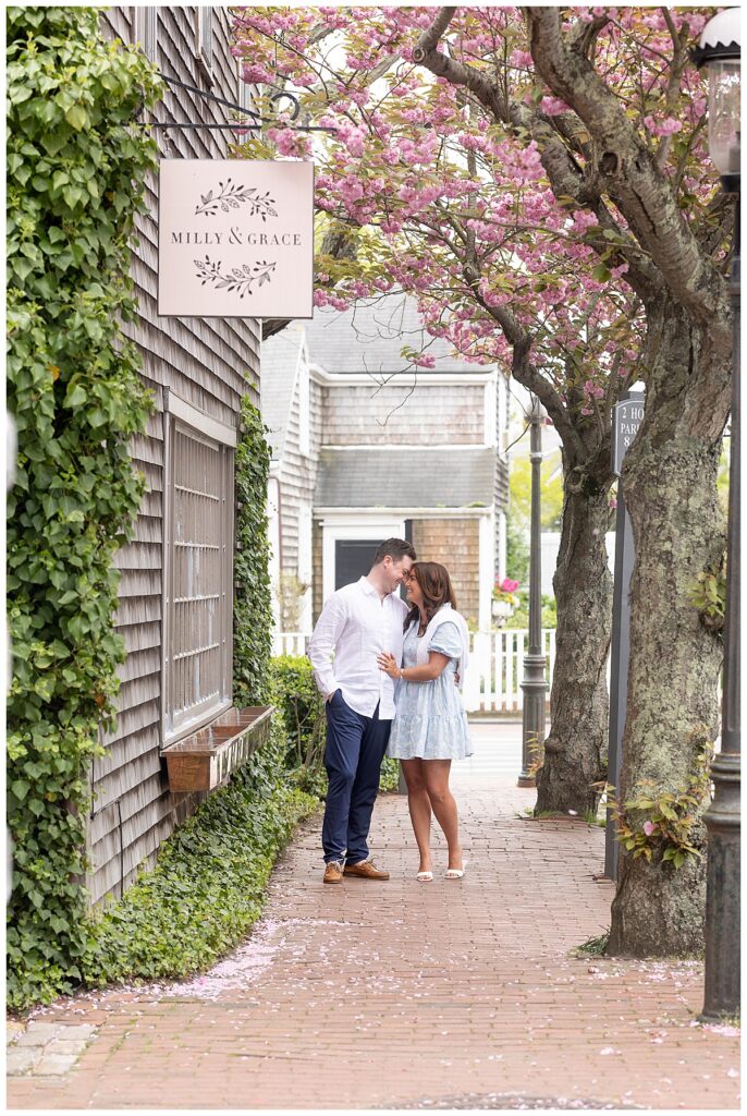 Couple stand under a pink blooming tree in downtown Nantucket during engagement pictures.