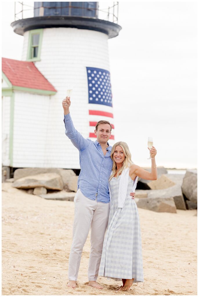 Couple raise a glass of champagne in the air during engagement pictures, while celebrating at Brant Point Lighthouse after a Nantucket Proposal.
