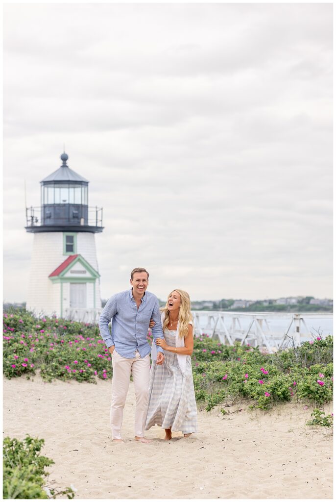 Couple walks hand in hand smiling and laughing by Brant Point Lighthouse in Nantucket after he proposed and they became engaged after his Nantucket Proposal.