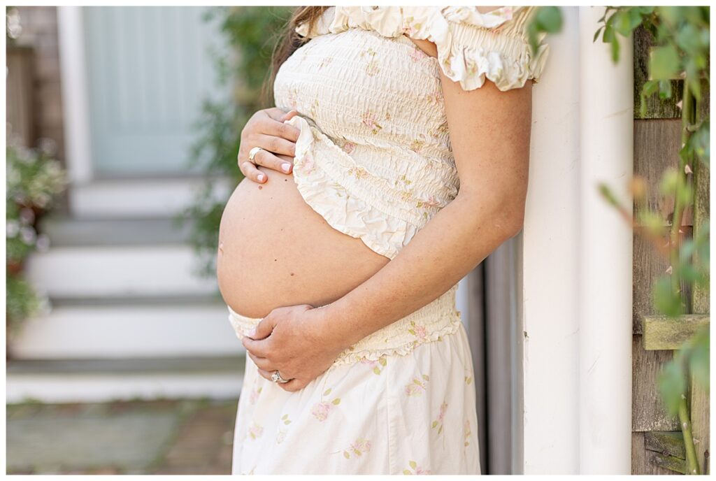 Woman shows pregnant belly off in Nantucket while having Maternity pictures taken and leaning against a gray shingled building.