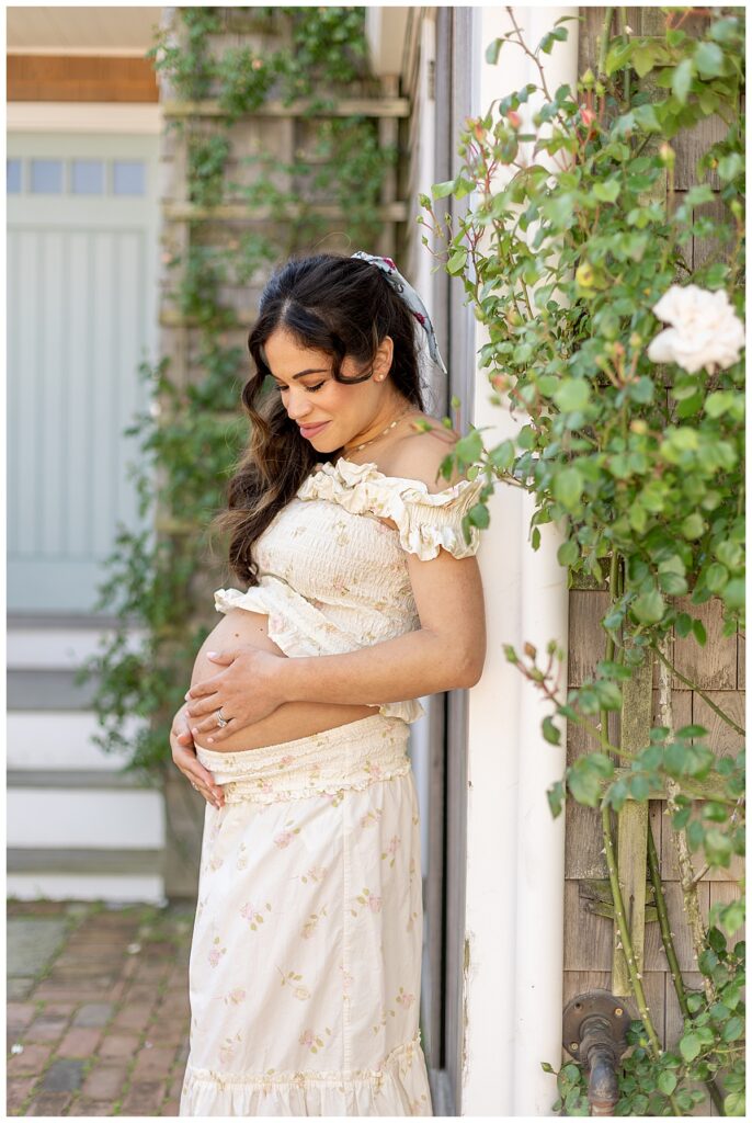 Woman looks lovingly at her belly while leaning against a gray shingled house with climbing roses in Nantucket during Maternity pictures.
