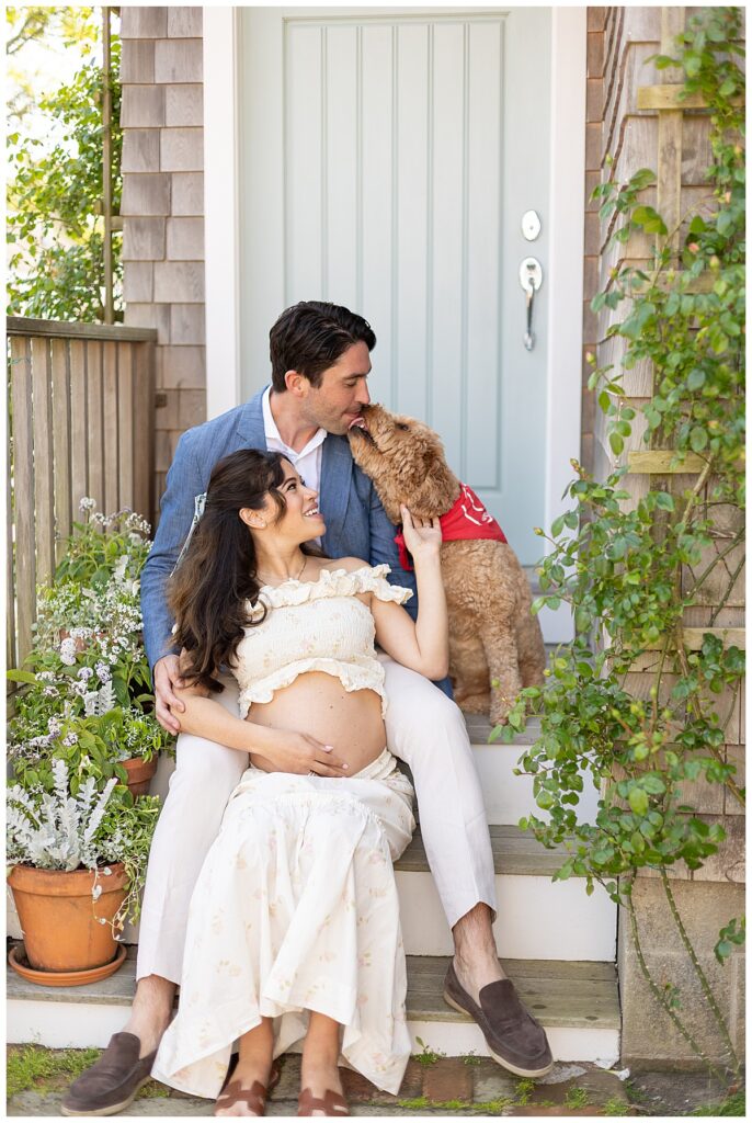 Husband, wife and goldendoodle smile and kiss one another while sitting on the steps of a Nantucket Boat Basin house while having Maternity pictures taken.