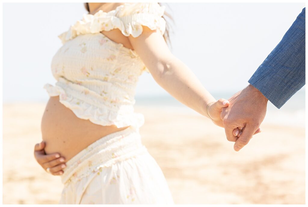Couple walks hand in hand on a beach in Nantucket during Maternity pictures. Her belly is showing and it is a beautiful summer day.