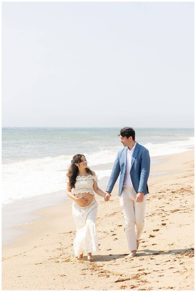 Husband and wife hold hands and stroll along the beach in Siasconset, Nantucket while having Maternity pictures taken.