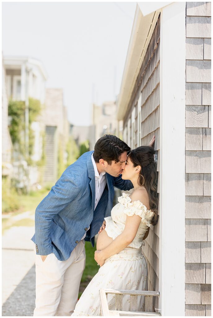 Husband and wife share a kiss while leaning against a gray shingled house along the Bluff Walk in Nantucket while having Maternity pictures taken.