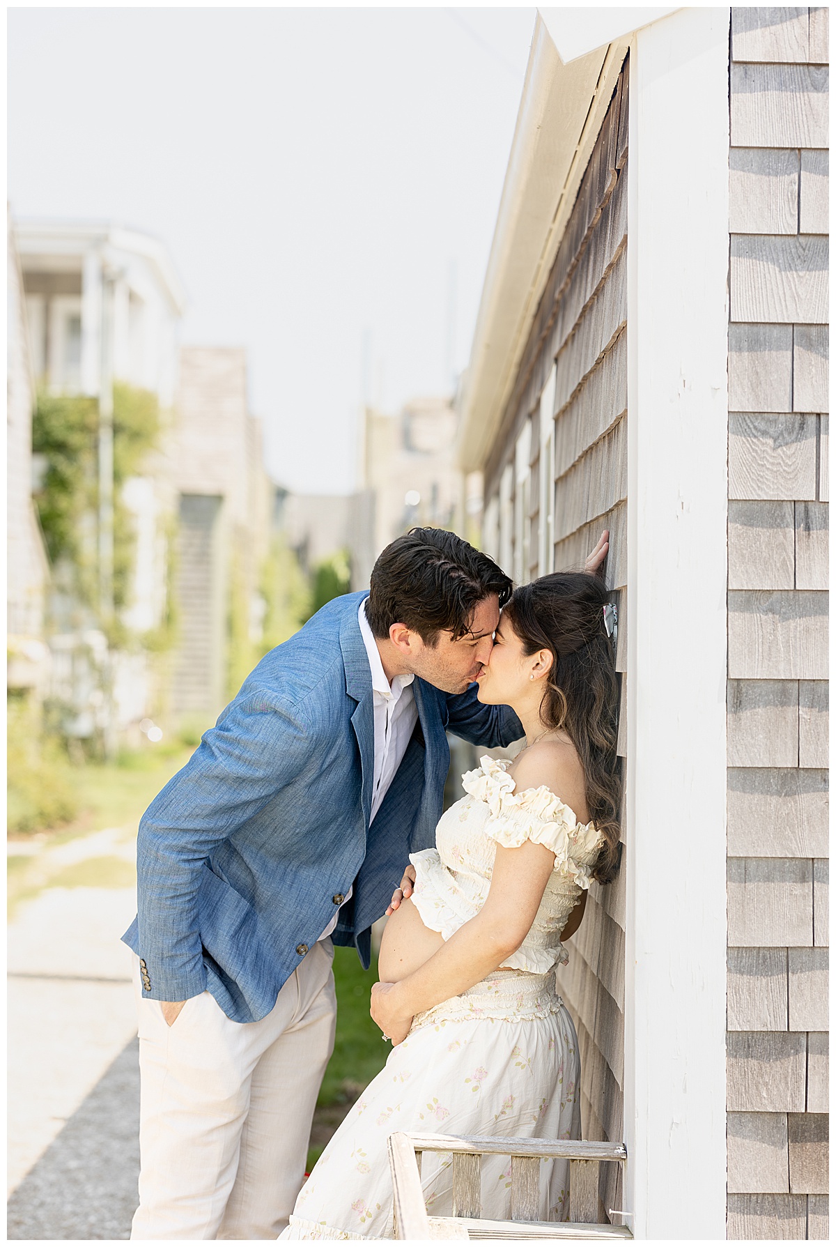 Husband and wife share a kiss while leaning against a gray shingled house along the Bluff Walk in Nantucket while having Maternity pictures taken.