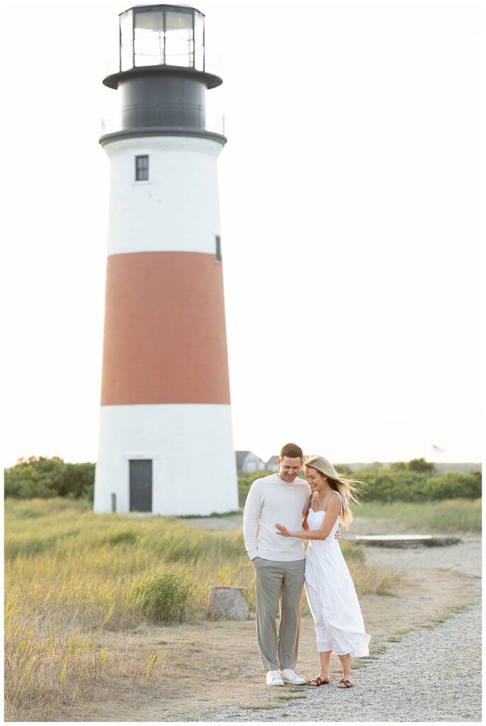 Man and woman stand in front of the Sankaty Head Lighthouse in Nantucket while the woman holds out her hand and looks at her new diamond ring after her Nantucket Proposal and Engagement.
