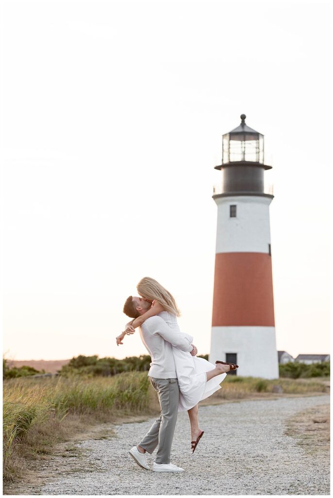 Man lifts woman up and kisses her with happy excitement in Nantucket at the Sankaty Head Lighthouse after his proposal and engagement on a warm July night at golden hour.