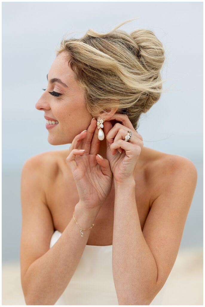 Bride shows off her bridal details at the beach, and gently shows the camera her earrings at her Chatham, Cape Cod wedding.