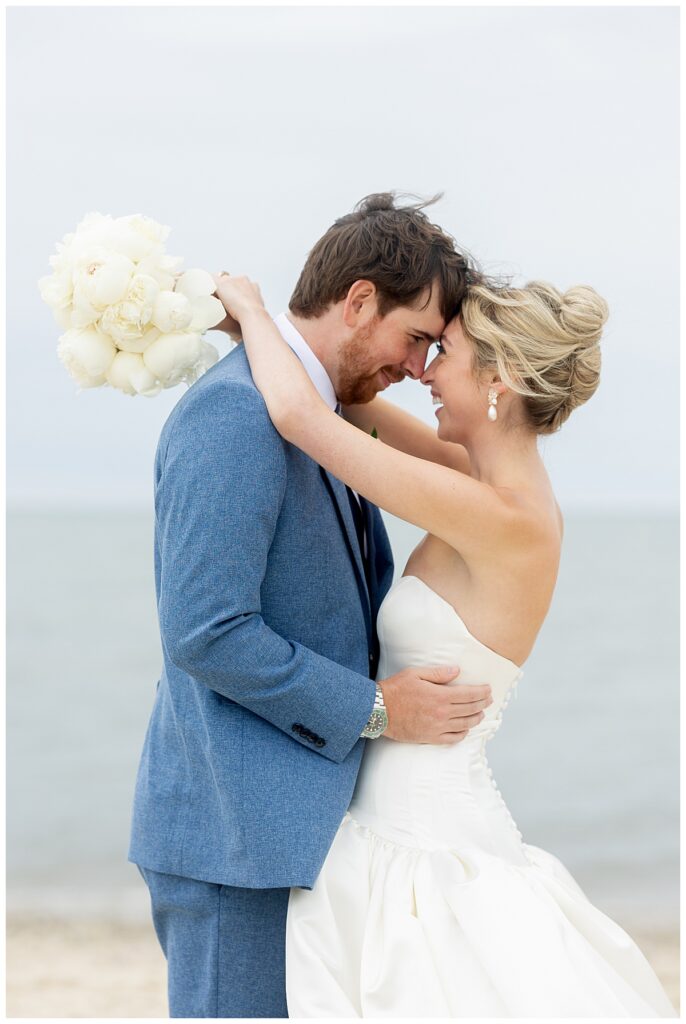 Bride drapes her arms around her Groom's head while they smile and touch foreheads on the beach at their Chatham, Cape Cod wedding.