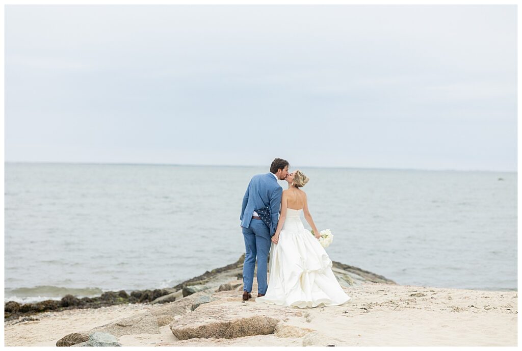 Bride and Groom walk along the jetty on the beach holding hands and share a kiss at their Chatham, Cape Cod wedding.