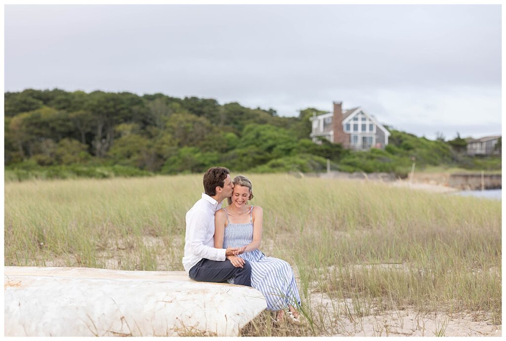 Man kisses woman on the temple, while sitting on an inside down canoe in the seagrass at the oceans edge after proposing to her in Chatham, Cape Cod, Massachusetts.