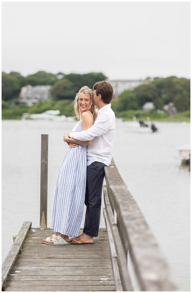 Woman laughing while man holds her back against his chest while leaning on the railing on a dock near the ocean in Chatham, Cape Cod, Massachusetts.