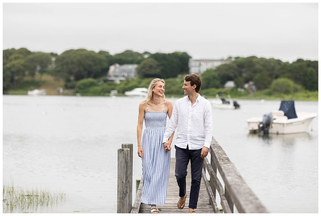 Couple walk and smile at one another while holding hands during engagement pictures in Chatham, Cape Cod, Massachusetts.