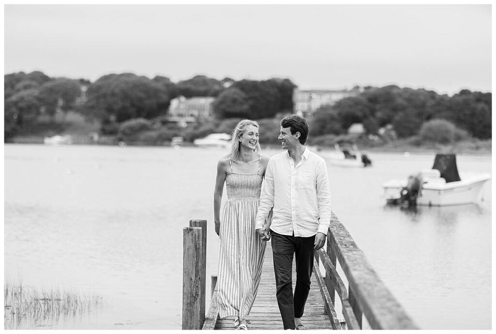 Black and white image of a couple walking hand and hand on a dock after getting engaged in Chatham, Cape Cod, Massachusetts.