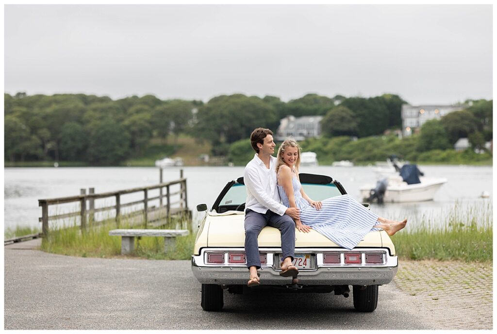 Man and woman hold hands and look at a ring on the woman's hand while sitting on a pale yellow vintage car during engagement pictures in Chatham, Cape Cod, Massachusetts.