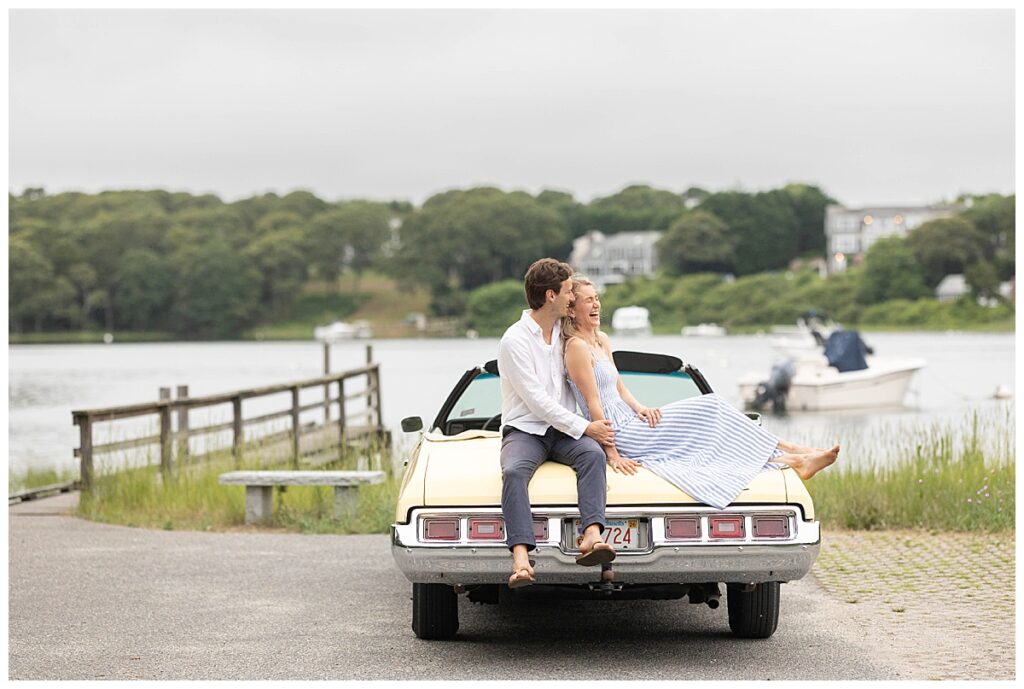 Couple laughs and sits on a canary yellow vintage car during engagement pictures in Chatham, Cape Cod, Massachusetts.
