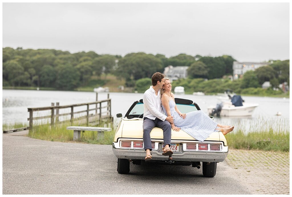 Man and woman sit on canary yellow vintage car and laugh during engagement pictures in Chatham, Cape Cod, Massachusetts.