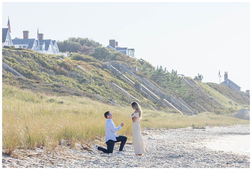 Man gets down on one knee on Steps Beach in Nantucket while proposing to his girlfriend wearing a pale yellow dress.