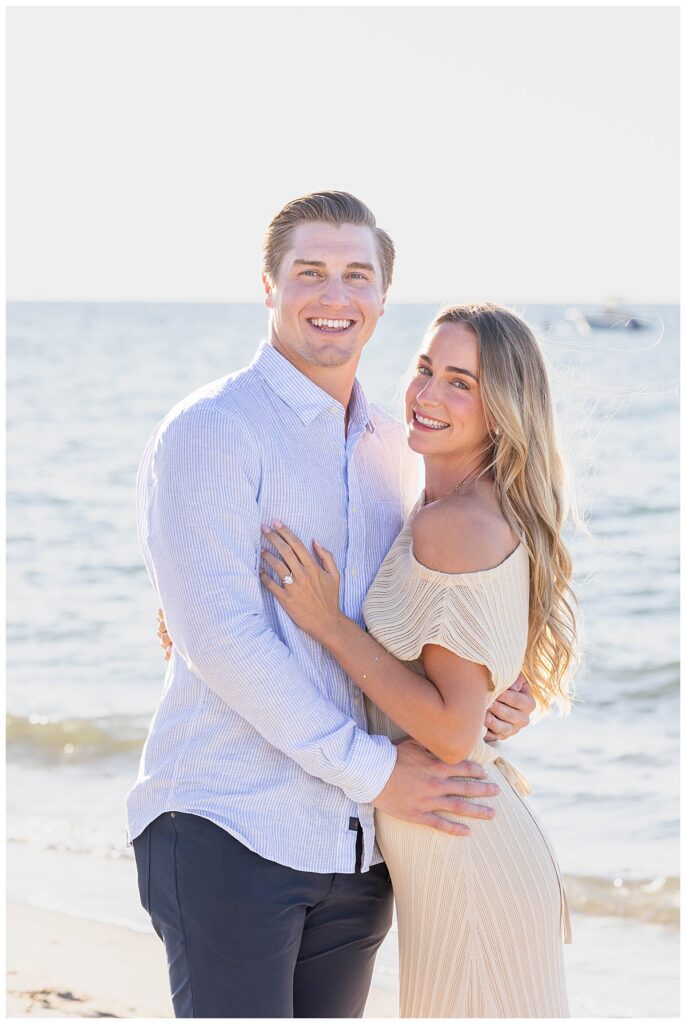Man and woman smile at the camera while standing on Steps Beach in Nantucket after his proposal and them getting engaged.