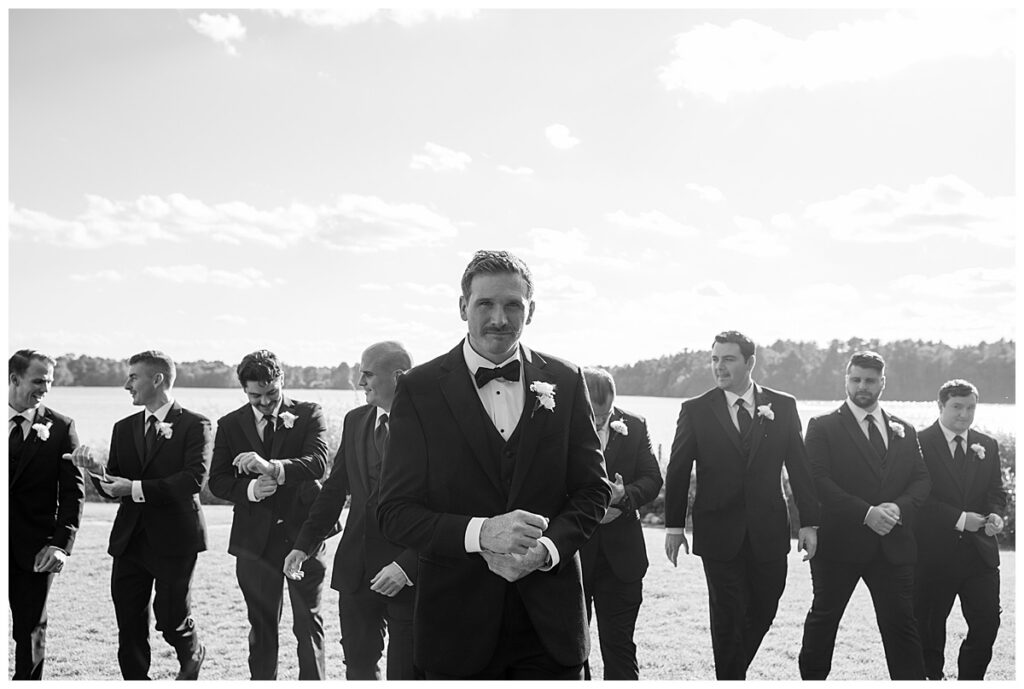 Groom and Groomsmen walk on lawn in black and white photo while adjusting cuff links and ties at The Lakehouse, a Sapphire Event Group venue, in Halifax, Cape Cod, Massachusetts.