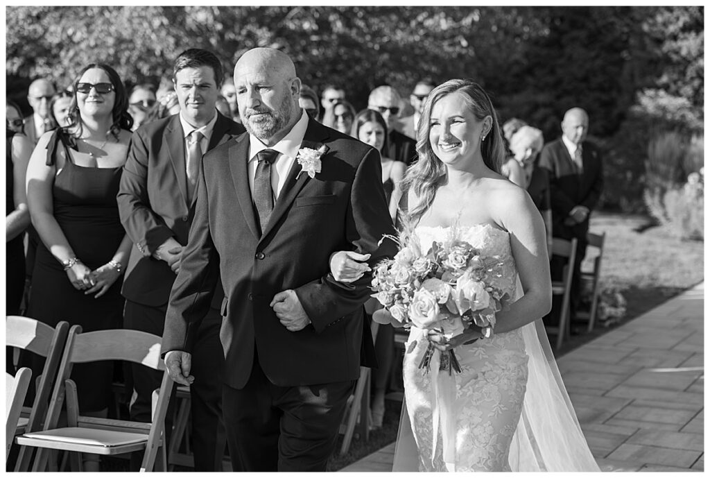 Black and white image of a father walking his daughter down the aisle at The Lakehouse, a Sapphire Event Group venue in Halifax, Cape Cod, Massachusetts.