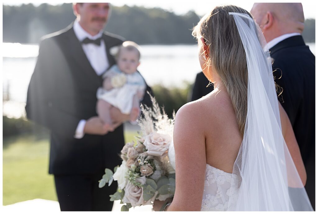 Bride is walked down the aisle as she approaches her groom holding their baby girl at the beginning of their ceremony at their wedding at The Lakehouse, a Sapphire Event Group venue, in Halifax, Cape Cod, Massachusetts.