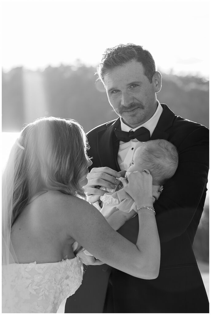 Groom looks at the camera while holding his baby daughter as his wife just walked down the aisle at The Lakehouse, a Sapphire Event Group venue in Halifax, Cape Cod, Massachusetts.