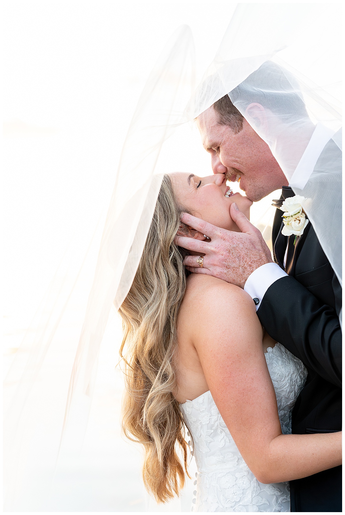 Groom cups his brides face as he goes into kiss her under her veil while standing on a dock at sunset at The Lakehouse, a Sapphire Event Group venue in Halifax, Cape Cod, Massachusetts.
