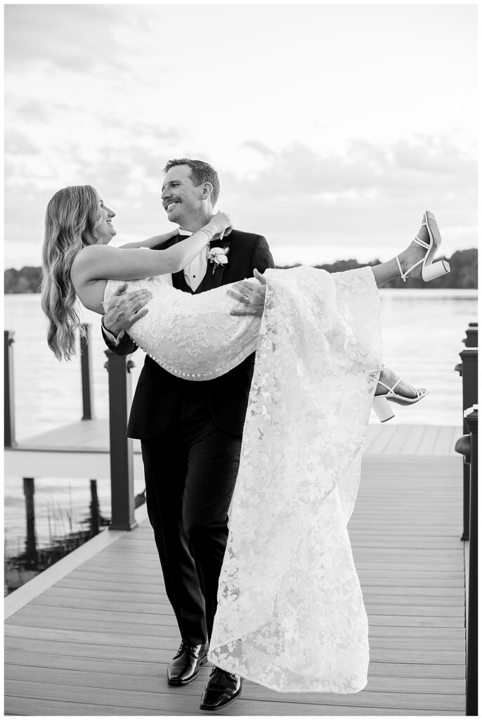 Black and white image of a groom carrying his bride off the dock to the grass at The Lakehouse, a Sapphire Event Group venue in Halifax, Cape Cod, Massachusetts.