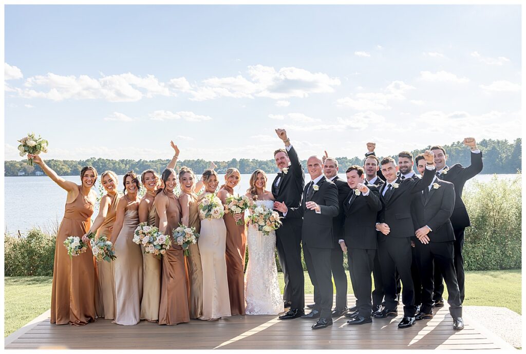Wedding party stands on in front of the water on a sunny day in September at The Lakehouse, a Sapphire Event Group venue.
