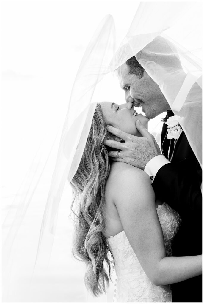 Black and white image of a groom kissing bride under a veil at their wedding in Halifax, Cape Cod, Massachusetts at sunset.