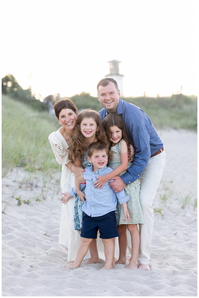 Family of five do a group hug and smile for the camera in front of the Chatham Lighthouse beach at sunset in Chatham, Cape Cod, Massachusetts.
