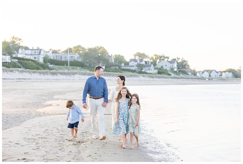 Husband and wife hold hands and smile and laugh at one another while walking along the shoreline at Chatham Lighthouse Beach at sunset in Chatham, Cape Cod, Massachusetts.