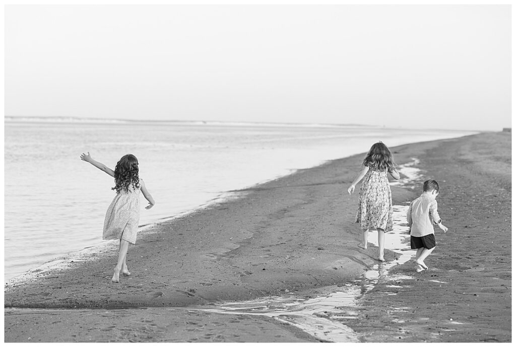 Black and white image of two young girls and one young boy running alone the shoreline at Chatham Lighthouse Beach at sunset.