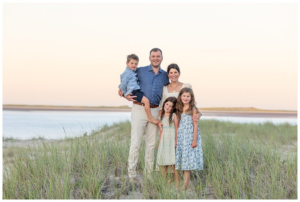 Family smiles for the camera while standing in seagrass on top of a sand dune overlooking the ocean at Chatham Lighthouse beach in Chatham, Cape Cod, Massachusetts.