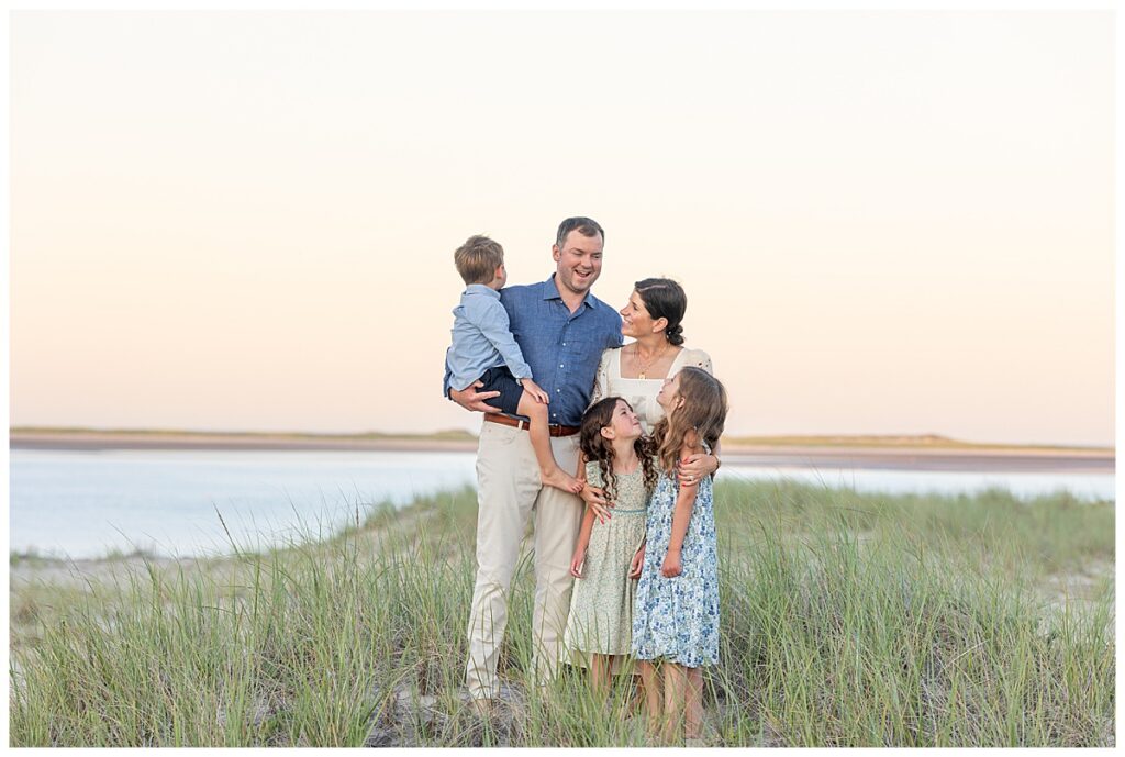 Family stands on a sand dune in the seagrass overlooking the ocean while looking at one another at Chatham Lighthouse beach in Chatham, Cape Cod, Massachusetts.