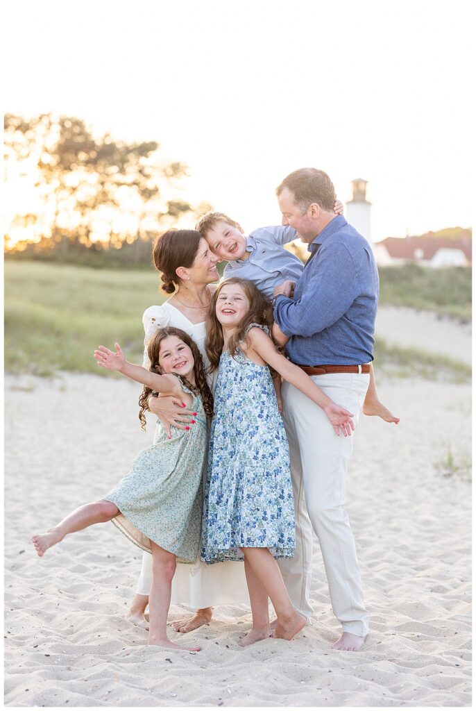 Family stands in front of the lighthouse and smiles and laughs while hugging one another at Chatham Lighthouse beach in Chatham, Cape Cod, Massachusetts.