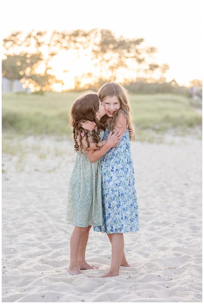 Sisters hug and the little sister kisses big sister on the cheek at sunset at Chatham Lighthouse beach in Chatham, Cape Cod, Massachusetts.