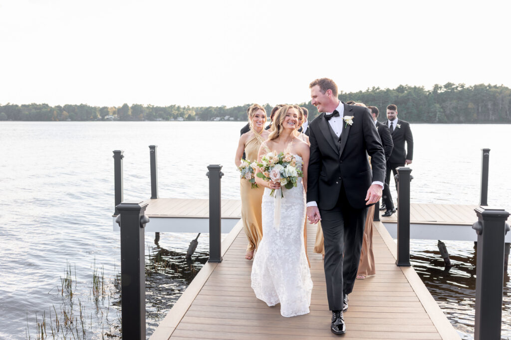 Bride and Groom walk hand in hand with bridal party following behind them as they exit the dock near the water at The Lakehouse, a Sapphire Event Group venue in Halifax, Cape Cod, Massachusetts.